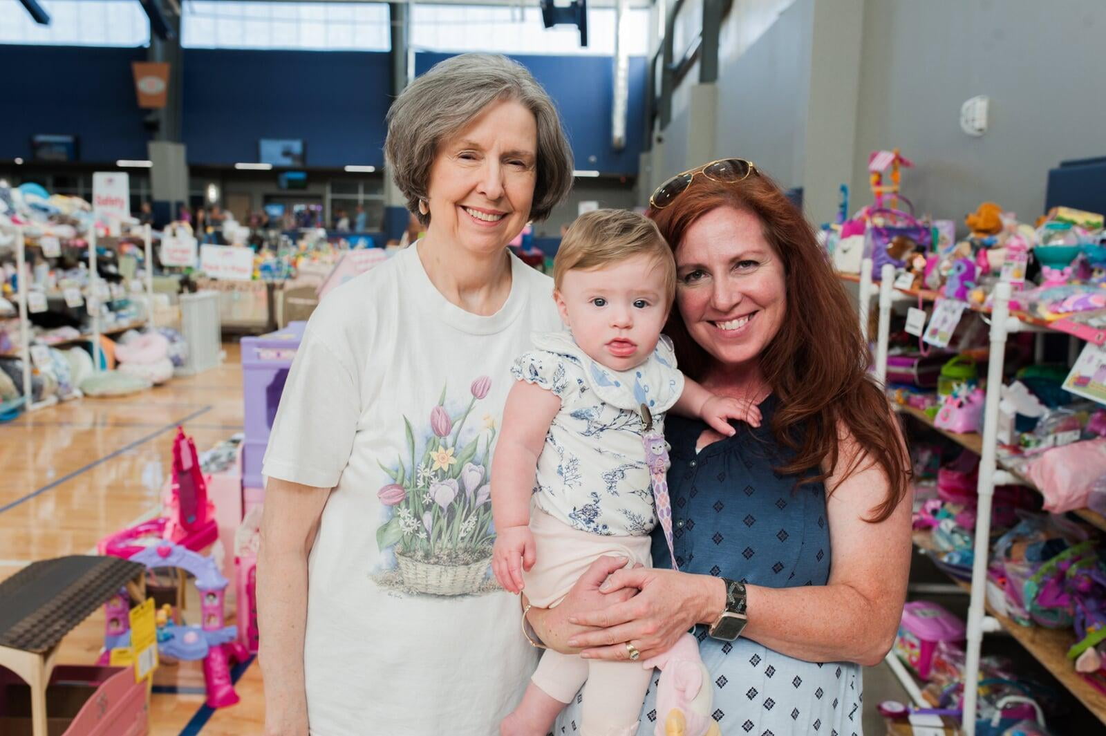 A young JBF shopper mom is holding her toddler while standing next to her child's grandmother at their local JBF sale. The background has racks of colorful toys.