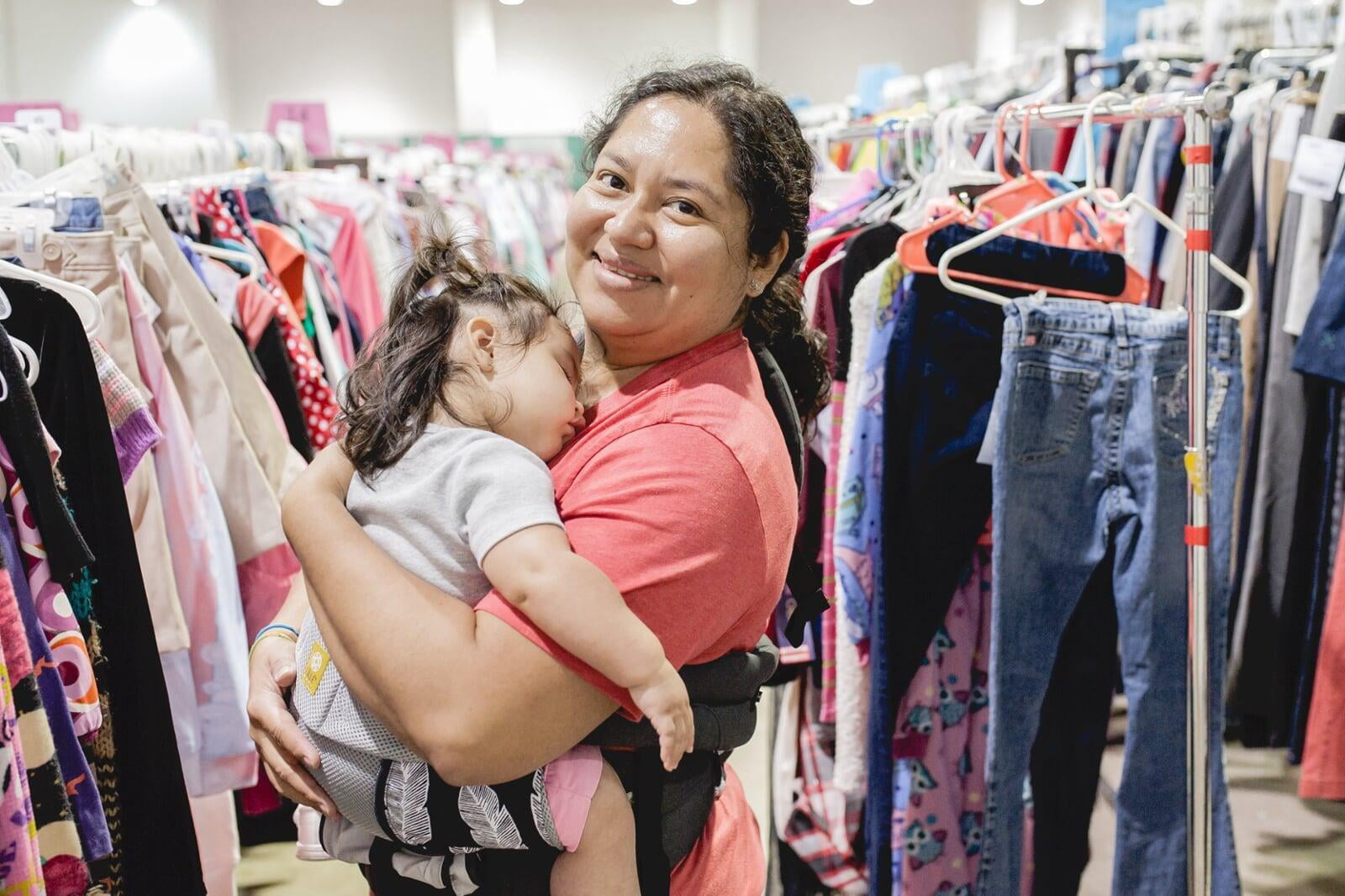 A mother is smiling while holding her sleeping toddler as she browses the racks at her JBF sale.