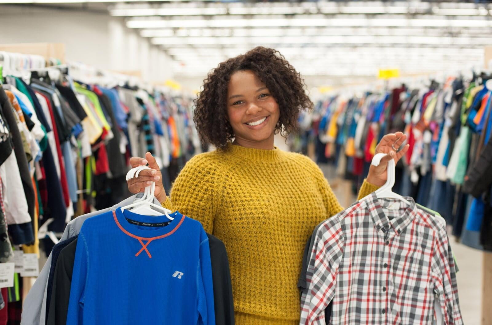 A women in a yellow sweater holds clothes on hangers in both hands. She is smiling.