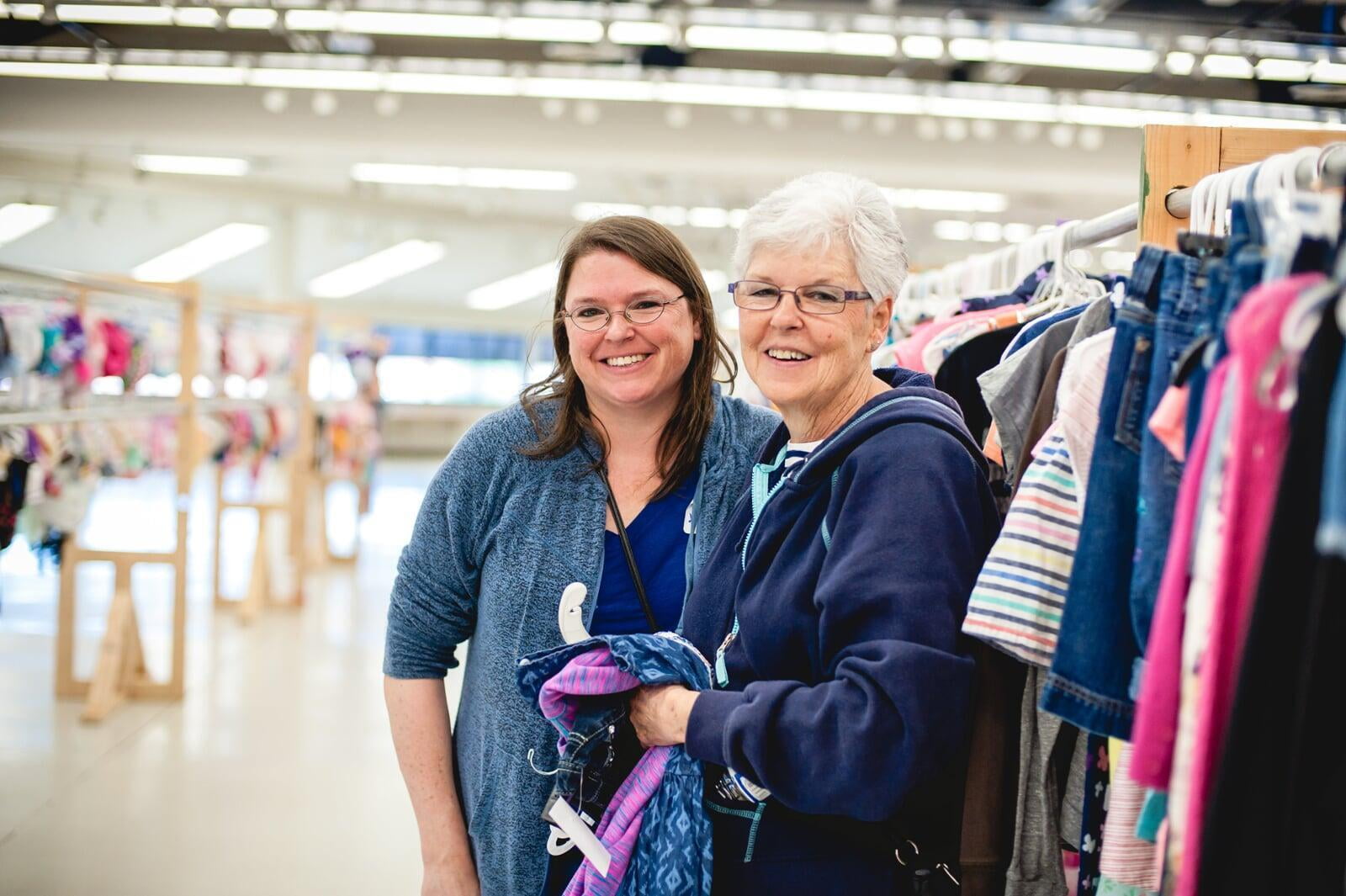 A mom and grandmother stand beside a rack of clothing at a JBF sale.