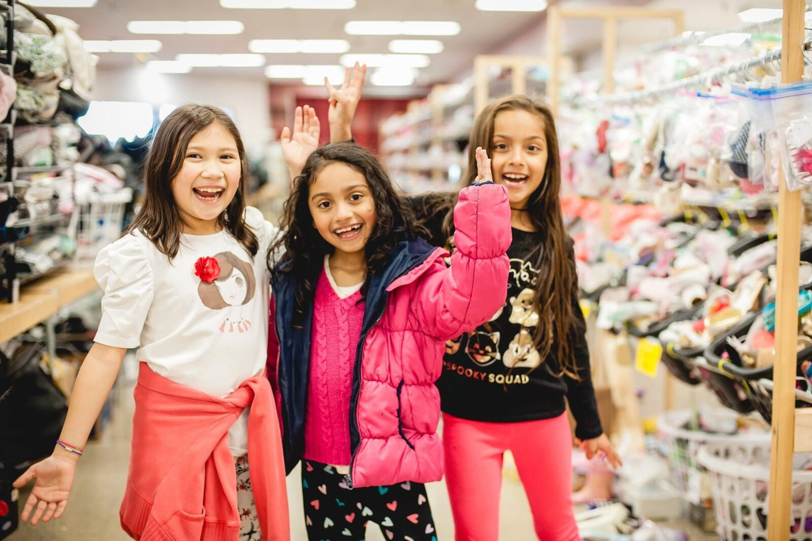 Three smiling girls stand between aisles at the JBF sale.