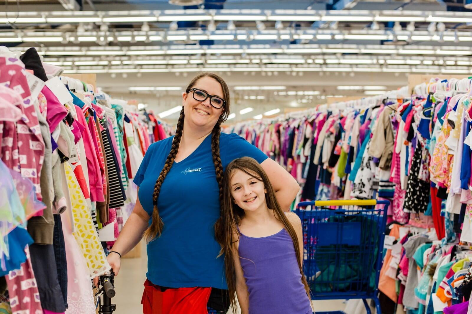 An employee is shopping with her daughter and is smiling towards the camera surrounded by handing clothes.