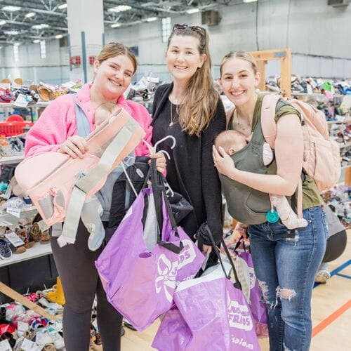 Three women are standing at a JBF sale. One woman is wearing her baby in a green holder. Another woman is holding two purple JBF bags. The third woman is holding a light pink baby carrier.