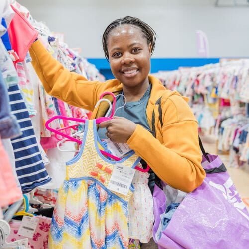 A woman in a yellow jacket stands in a clothing aisle at a JBF sale with one hand on the clothing rack and one hand holding several dresses on a hanger. She is smiling. 