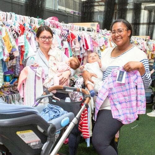Two friends are shopping together at a JBF sale. They are standing in a clothing aisle with their stroller. They are both holding their babies and smiling.