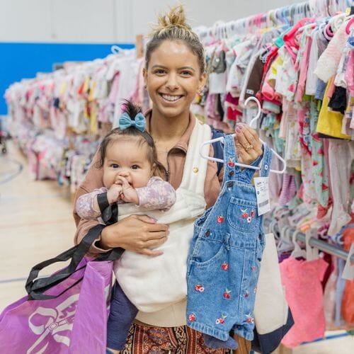 A woman stands in the clothing aisle of a JBF sale. She's wearing her baby in a white holder and holding a pair of blue overalls on a hanger. She is smiling and the baby is putting her hands together.